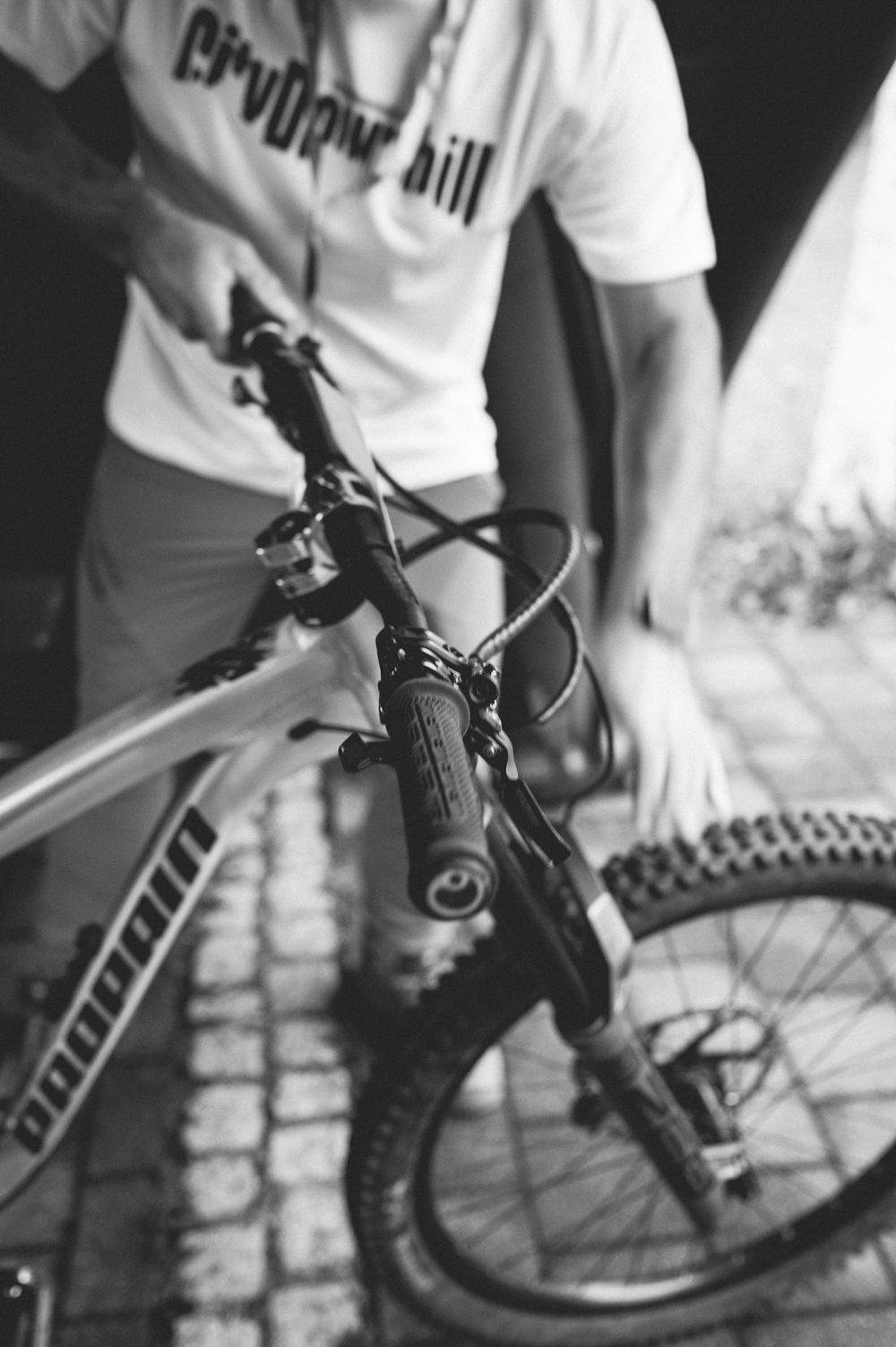 Black and white photo of a person in a white t-shirt standing over a mountain bike on cobblestone pavement.