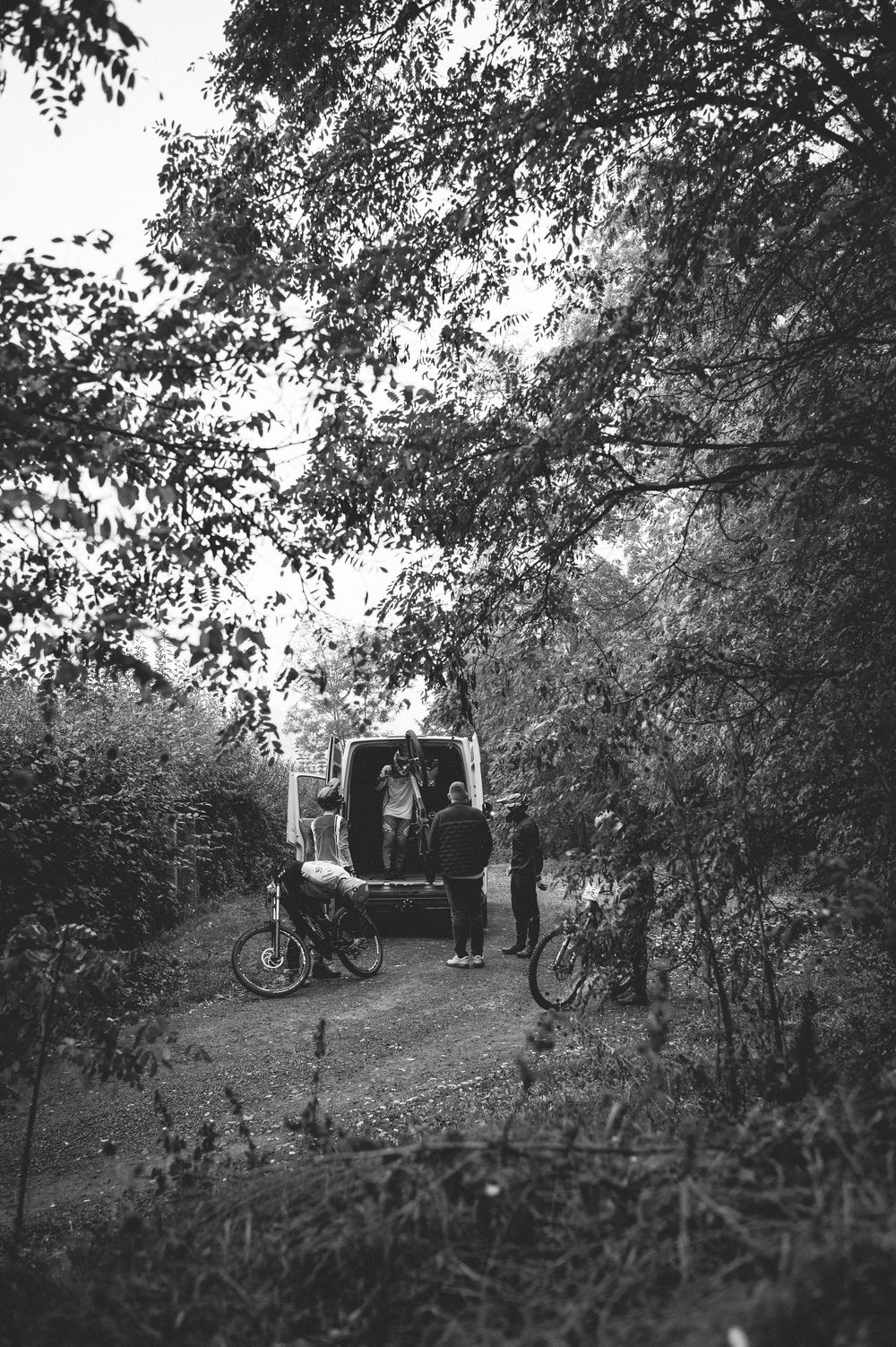 Black and white photo of cyclists unloading bikes from a van on a forest trail surrounded by lush trees.