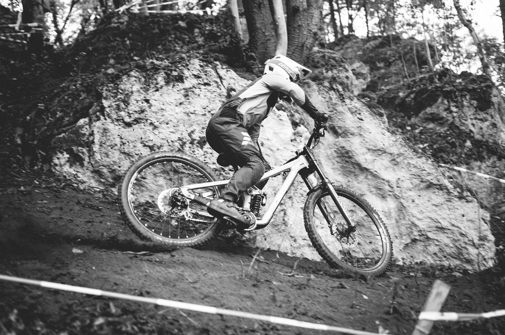 Black and white photo of a mountain biker racing downhill on a dirt trail through a wooded area.