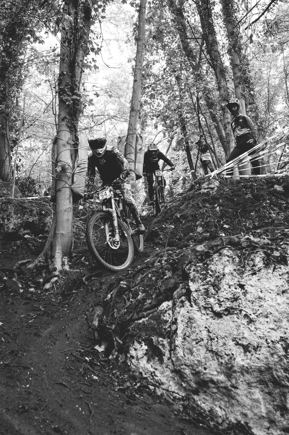 Black and white photo of mountain bikers racing downhill through a rocky forest trail during a competitive cycling event.