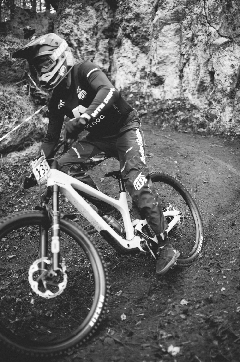 Black and white photo of a mountain biker in full gear racing downhill on a dirt trail during a competition.