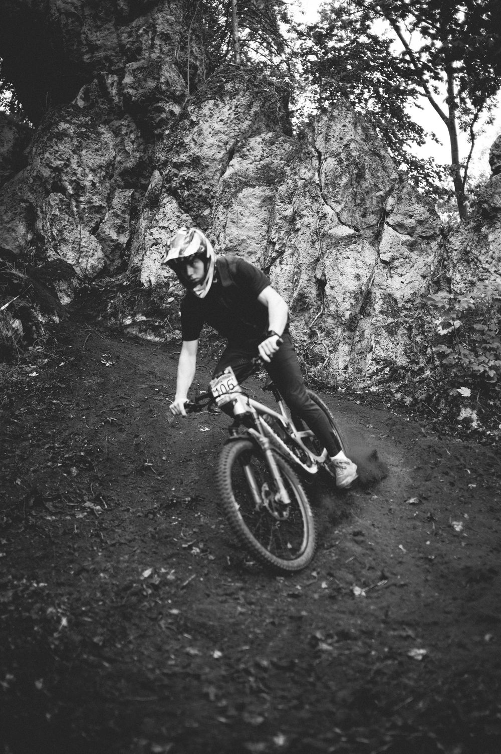 Mountain biker in helmet cornering on a dirt trail with rocky cliff background, black and white photo.