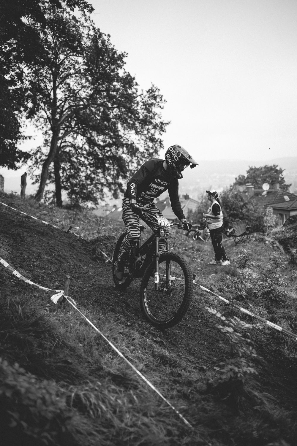 Black and white photo of a mountain biker racing downhill on a grassy slope wearing full protective gear.