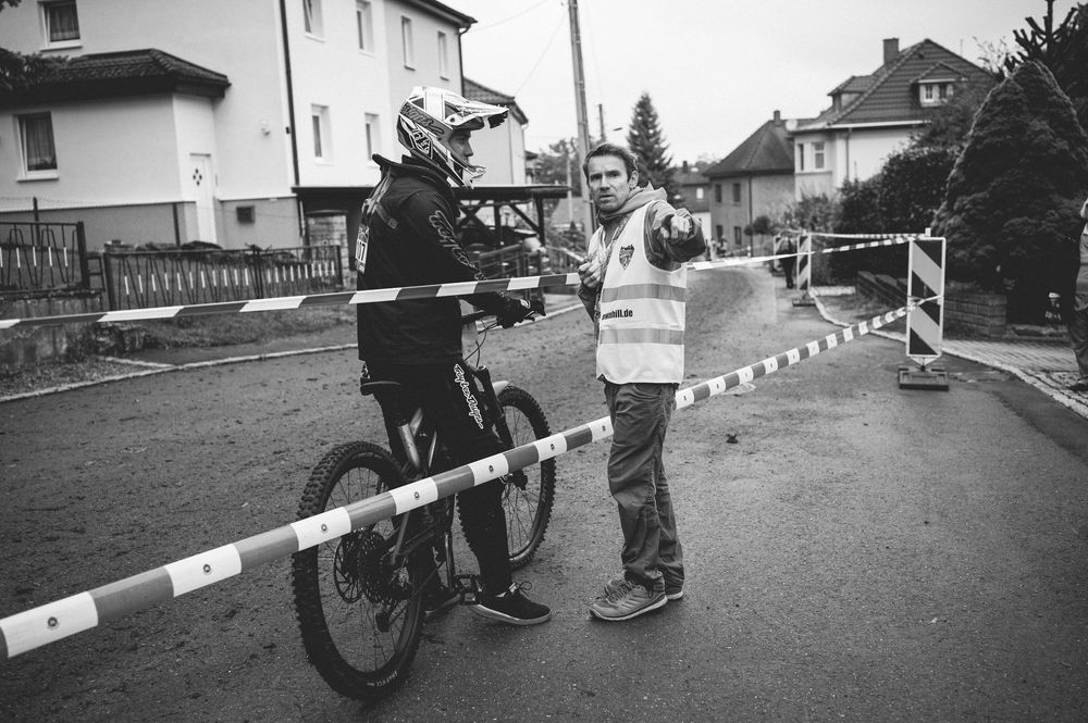 Black and white photo of a cyclist with helmet receiving directions from a race official near a barrier on a residential street.