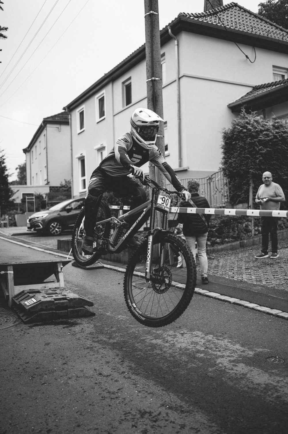 A helmeted rider performs a jump on a mountain bike during a street race in a residential area, in black and white.