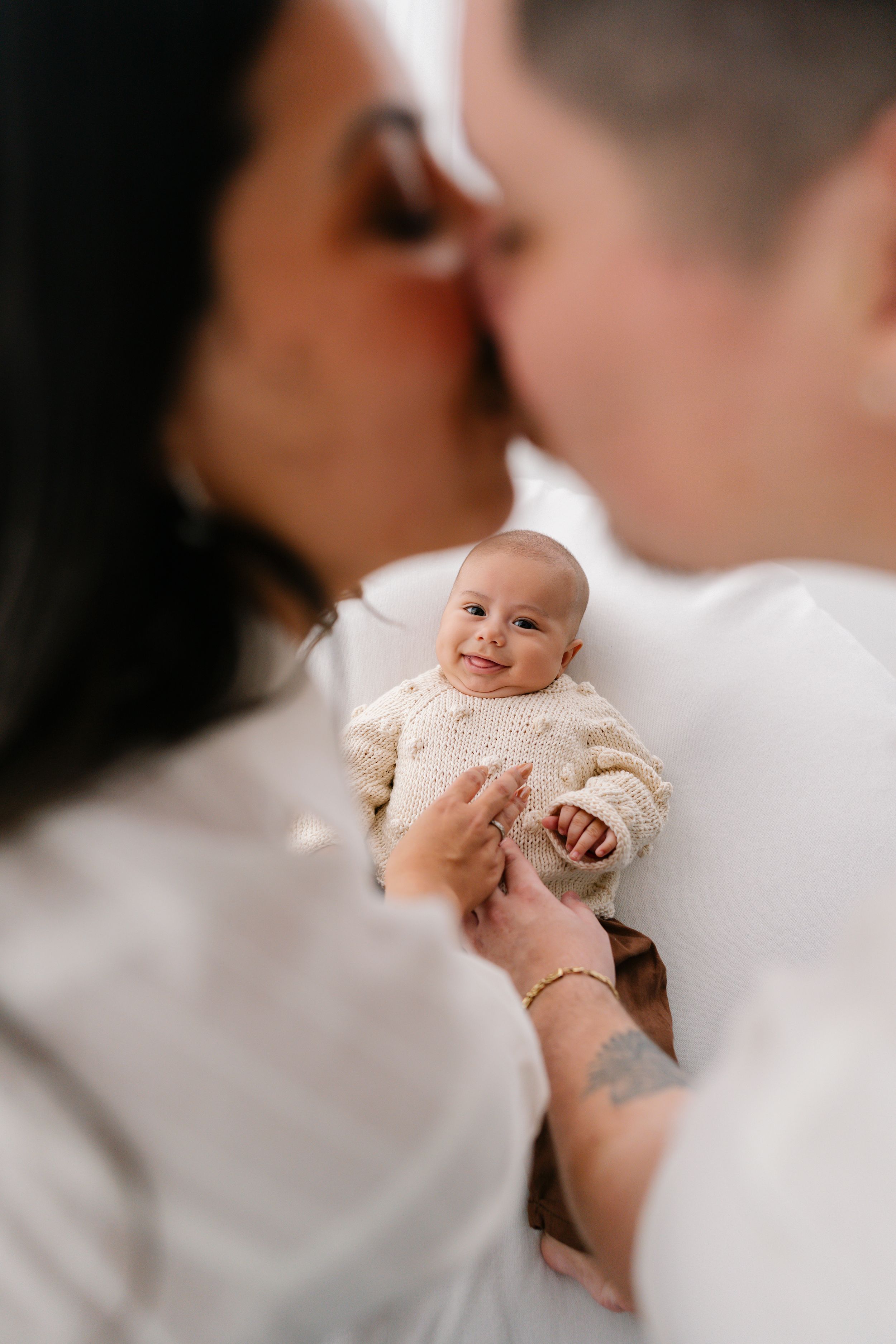 Two parents kissing while holding their smiling newborn baby dressed in a cream knit sweater.
