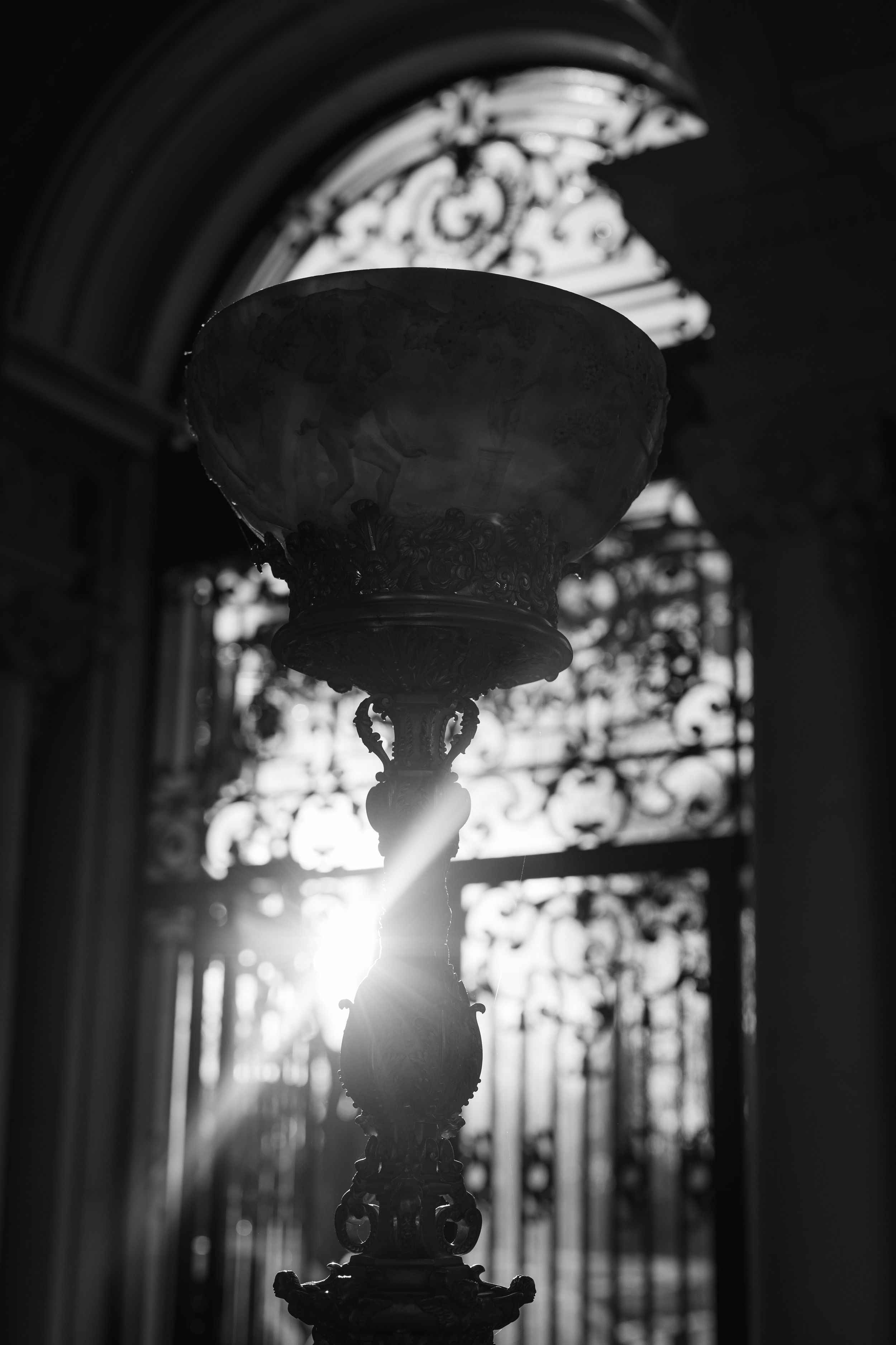 Black and white photo of ornate antique lamp with sunlight streaming through decorative iron gates in elegant interior.