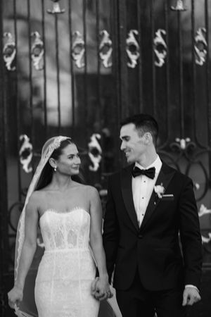 Black and white photo of a smiling bride in lace gown and groom in tuxedo holding hands before ornate iron gates.