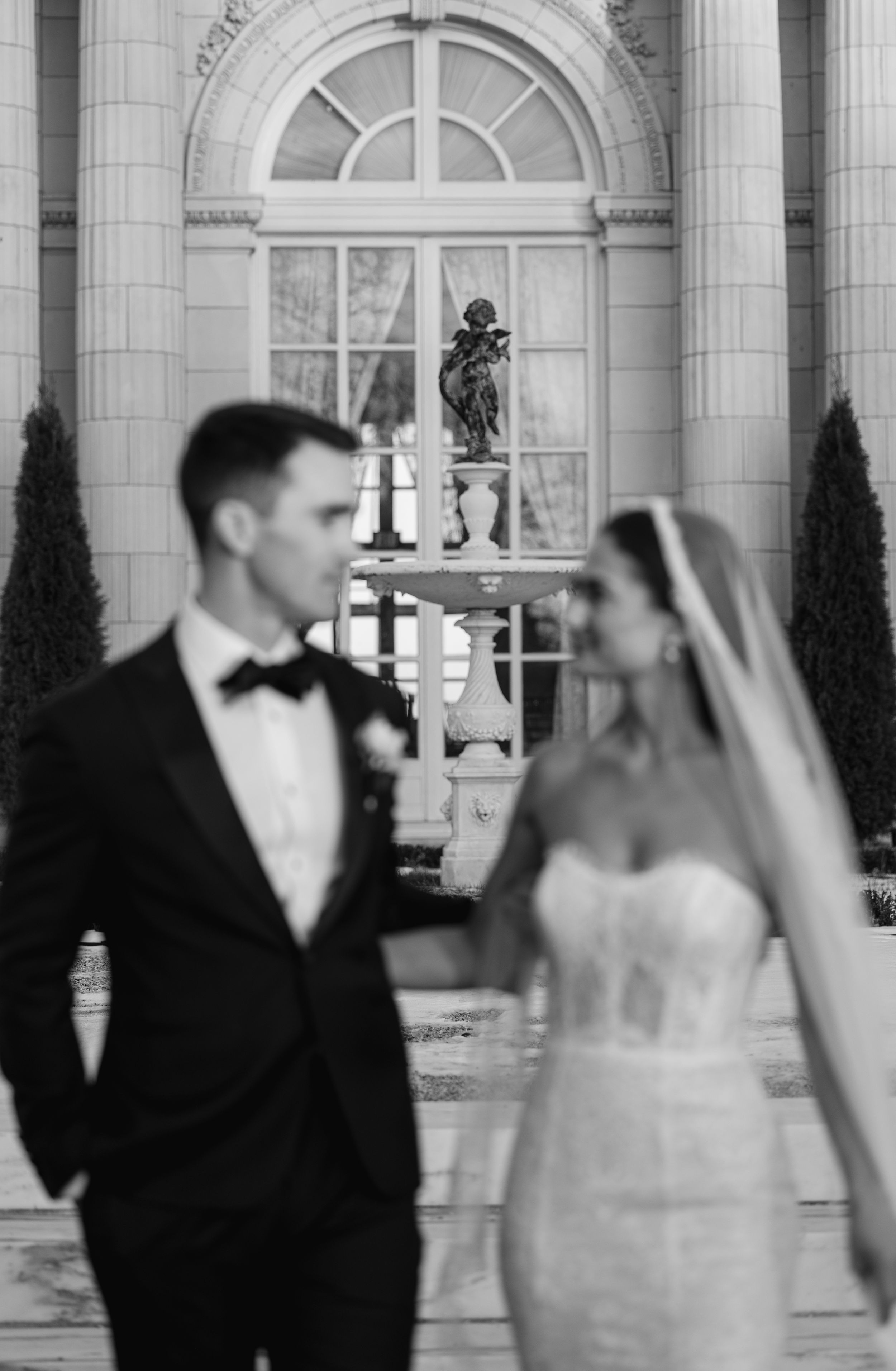 Black and white wedding photo of bride in strapless gown and groom in tuxedo posing before an elegant neoclassical building.