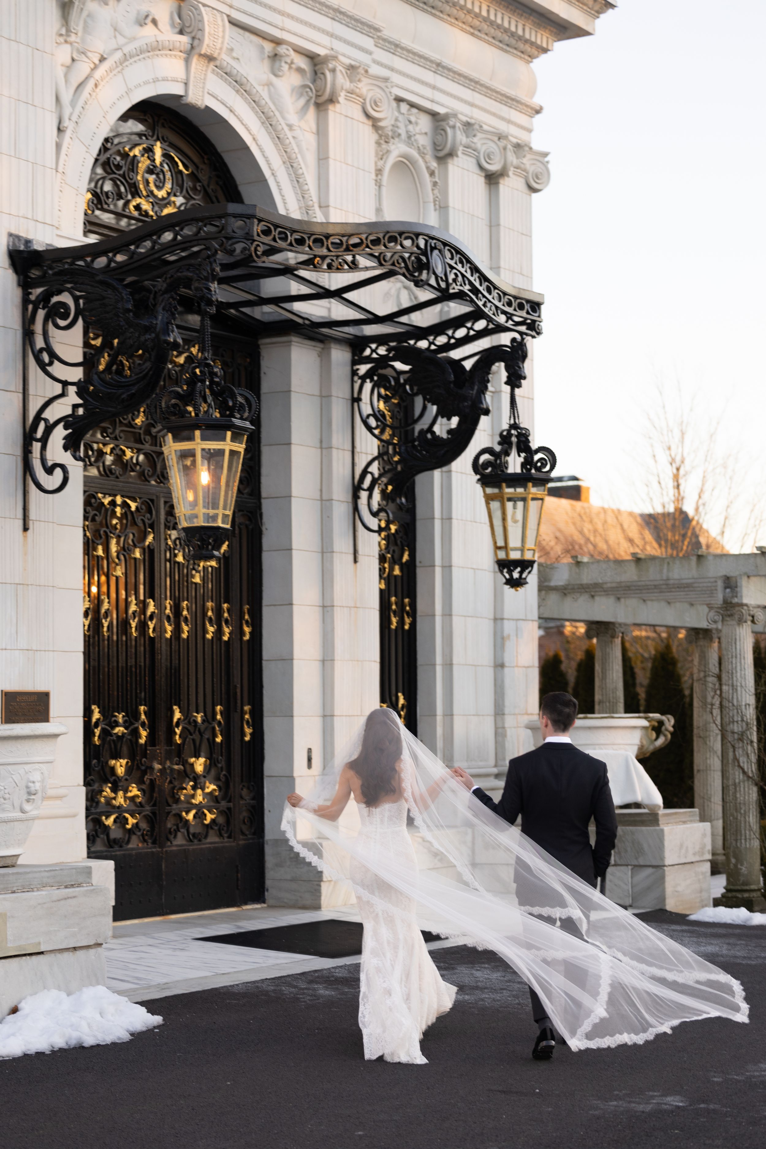 Bride and groom walking toward ornate black iron gates of an elegant white marble wedding venue.