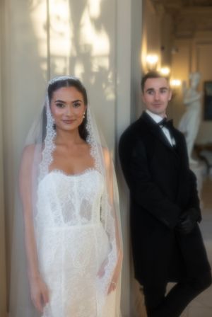 Bride in lace gown and veil poses with groom in black tuxedo at elegant wedding venue.