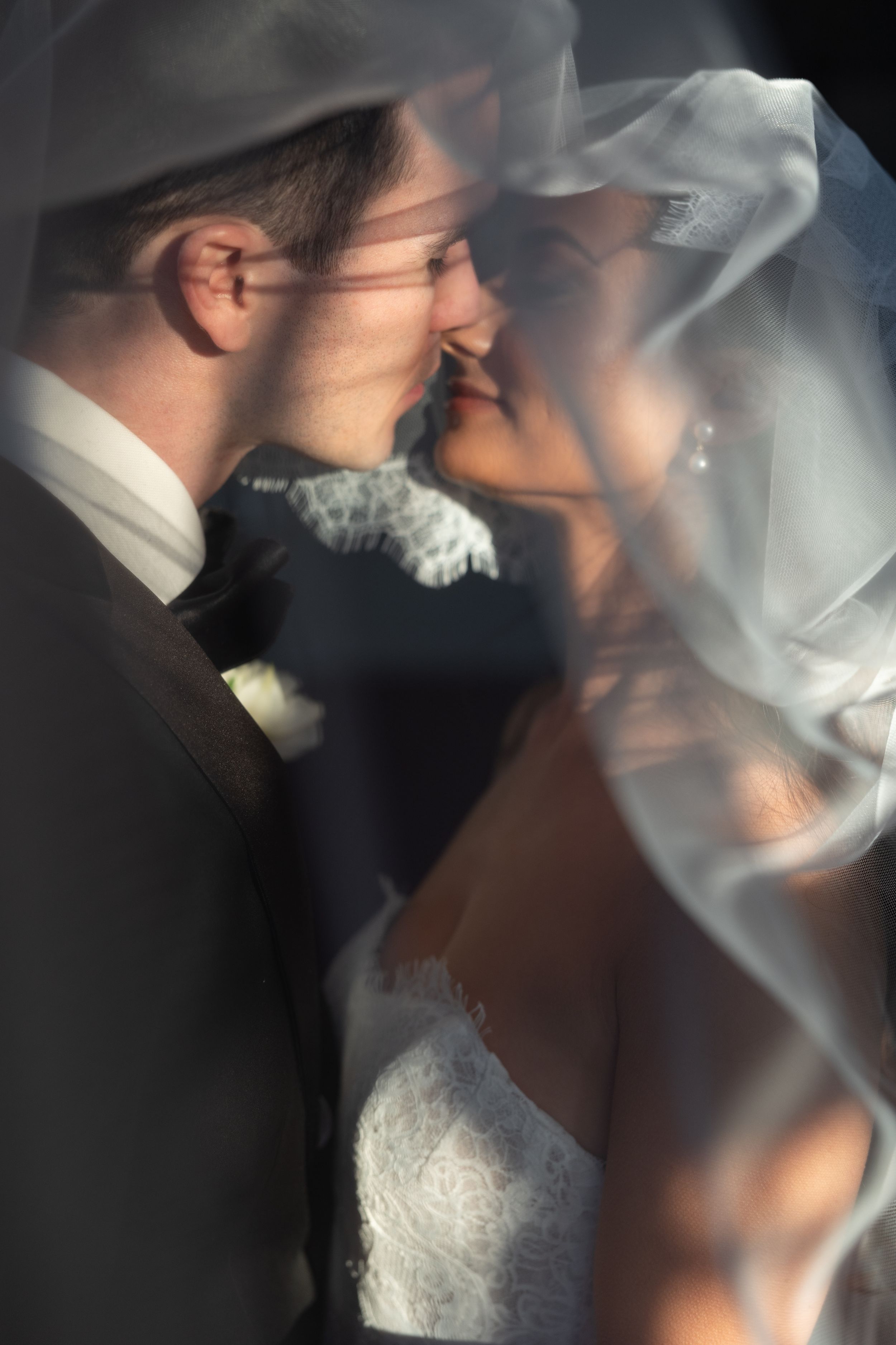 Bride and groom sharing an intimate moment under a flowing wedding veil, soft light highlighting their romantic embrace.