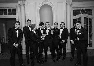 Black and white photo of groomsmen in tuxedos laughing together in an elegant ballroom setting.