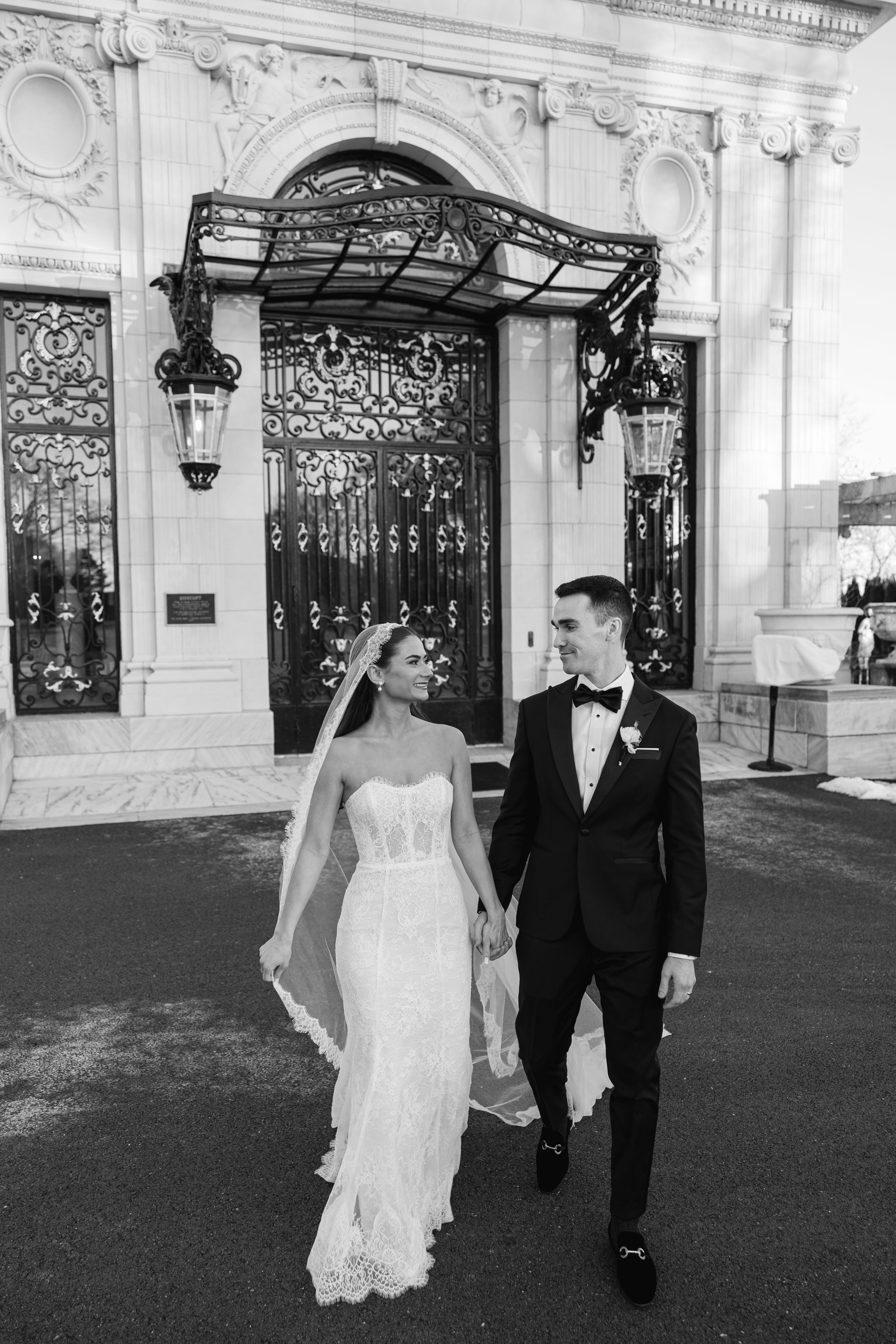 Black and white photo of bride and groom holding hands outside ornate building with iron gates.