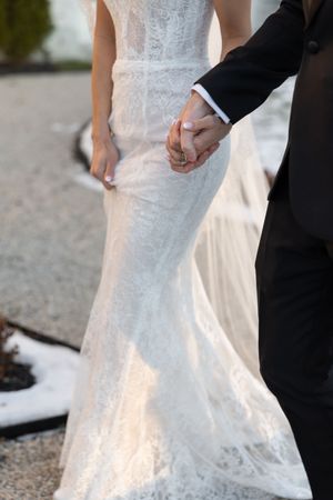 Bride in lace mermaid wedding gown holds hands with groom in black suit outdoors.