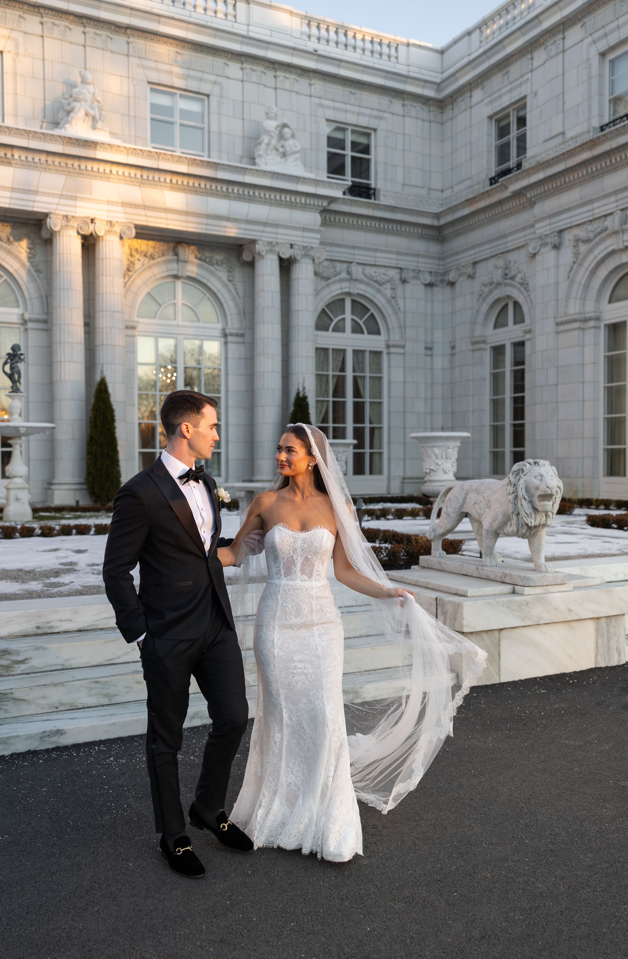 Bride and groom pose outside a grand neoclassical mansion with lion statues, her veil flowing in the breeze.