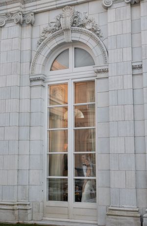Bride and groom visible through elegant arched French doors of a grand white stone building with ornate carved details.