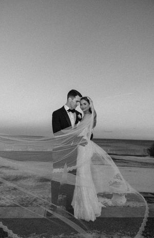 Romantic black and white wedding photo of couple embracing at sunset with flowing veil.