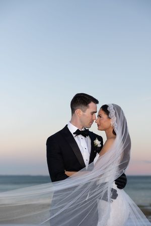 Bride and groom share a romantic moment by the ocean at dusk, veil flowing in wind.