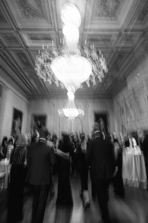 Blurred black and white photo of elegantly dressed guests mingling in a grand ballroom with crystal chandeliers.