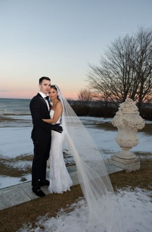 Bride in white lace gown and veil with groom in black tuxedo posing outdoors in snowy winter landscape at sunset.