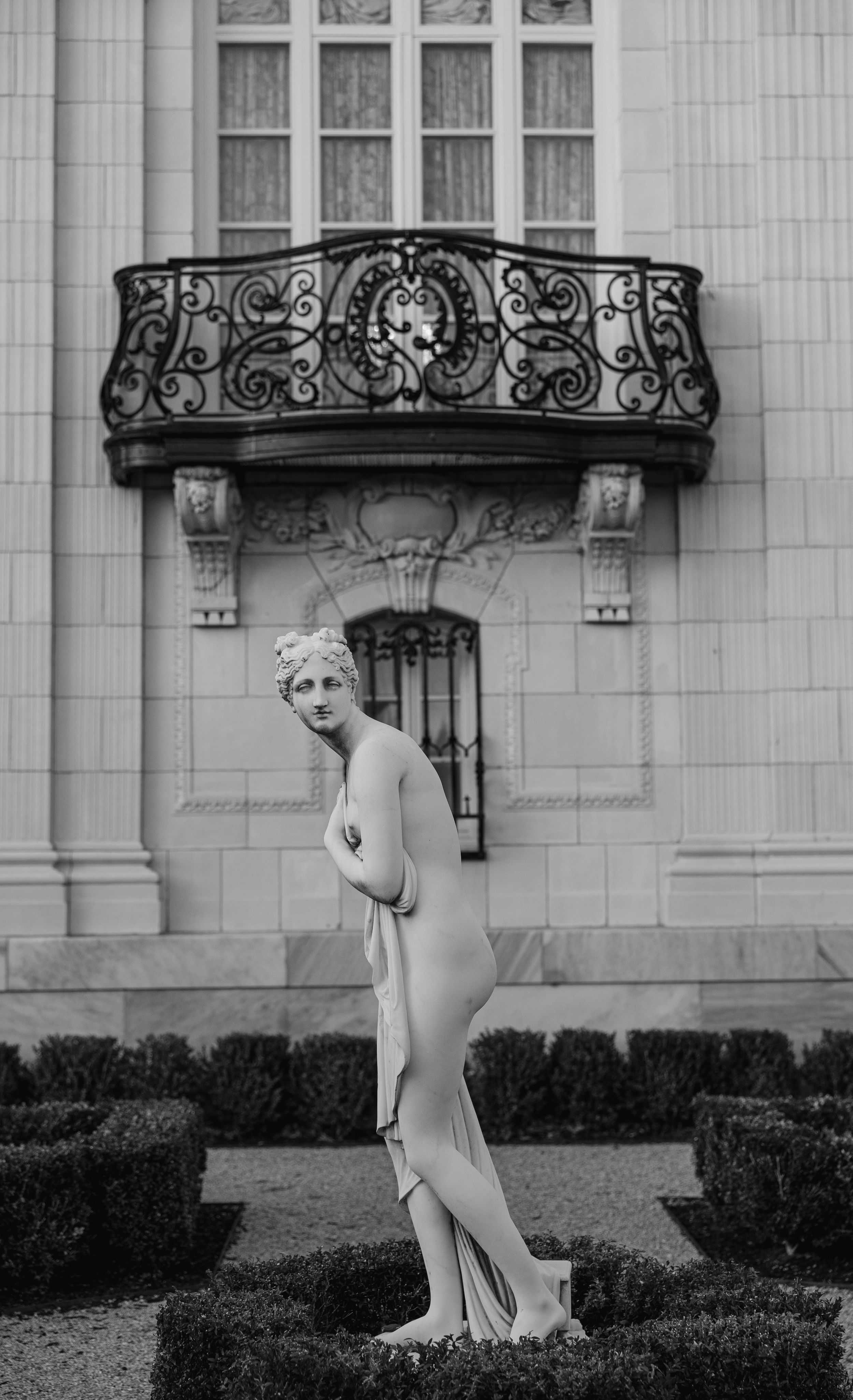 Black and white photo of a classical nude marble statue in a formal garden with ornate balcony.