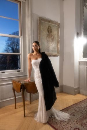 Bride in strapless white fitted gown holding black fur coat stands beside vintage portrait painting in elegant room.