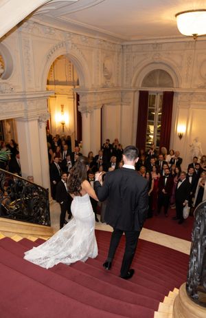 Bride and groom descend grand staircase before wedding guests in ornate ballroom.