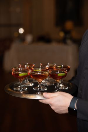 Waiter carrying silver tray of amber cocktails garnished with lime in coupe glasses at formal wedding reception.