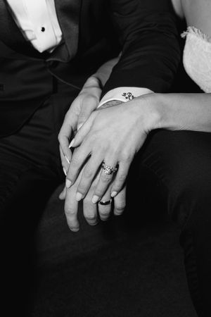 Close-up black and white photo of bride and groom holding hands, showing wedding and engagement rings.