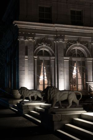 Stone lion statues flank illuminated neoclassical building entrance at night with arched windows and ornate columns.