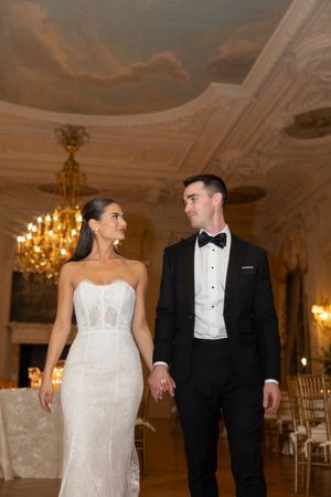 Bride and groom walk hand-in-hand through opulent ballroom with chandelier and murals.