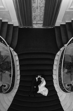 Aerial black and white view of bride and groom kissing at base of grand sweeping staircase.