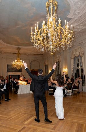 Newlyweds raise hands triumphantly on dance floor beneath ornate gold chandelier at wedding reception.