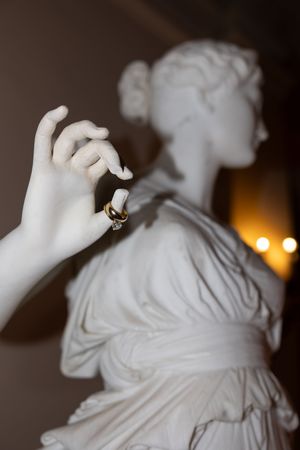 White marble classical statue holding gold wedding rings against a warm bokeh background.