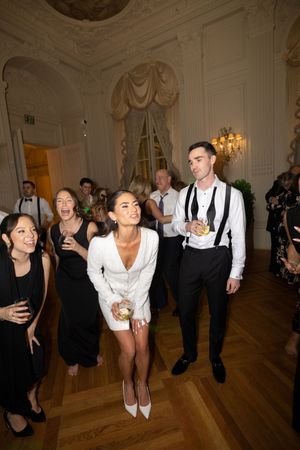 Guests dancing at elegant wedding reception in ornate ballroom with woman in white dress and man in suspenders.