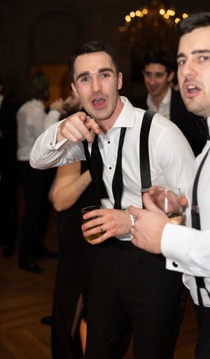 Young man in suspenders and white shirt pointing at camera during formal party event.