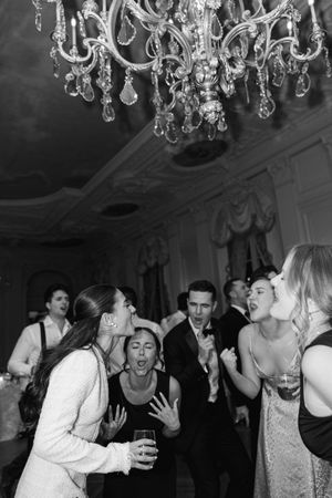Bride and guests sing and dance joyfully under crystal chandelier at wedding reception.