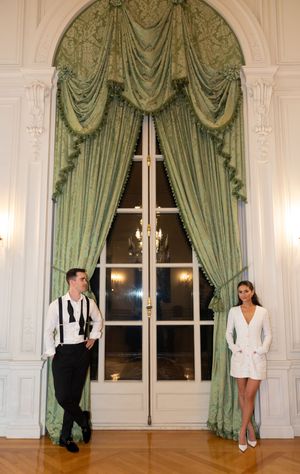 Couple posing by tall French doors with green damask drapes in a classic white ballroom.