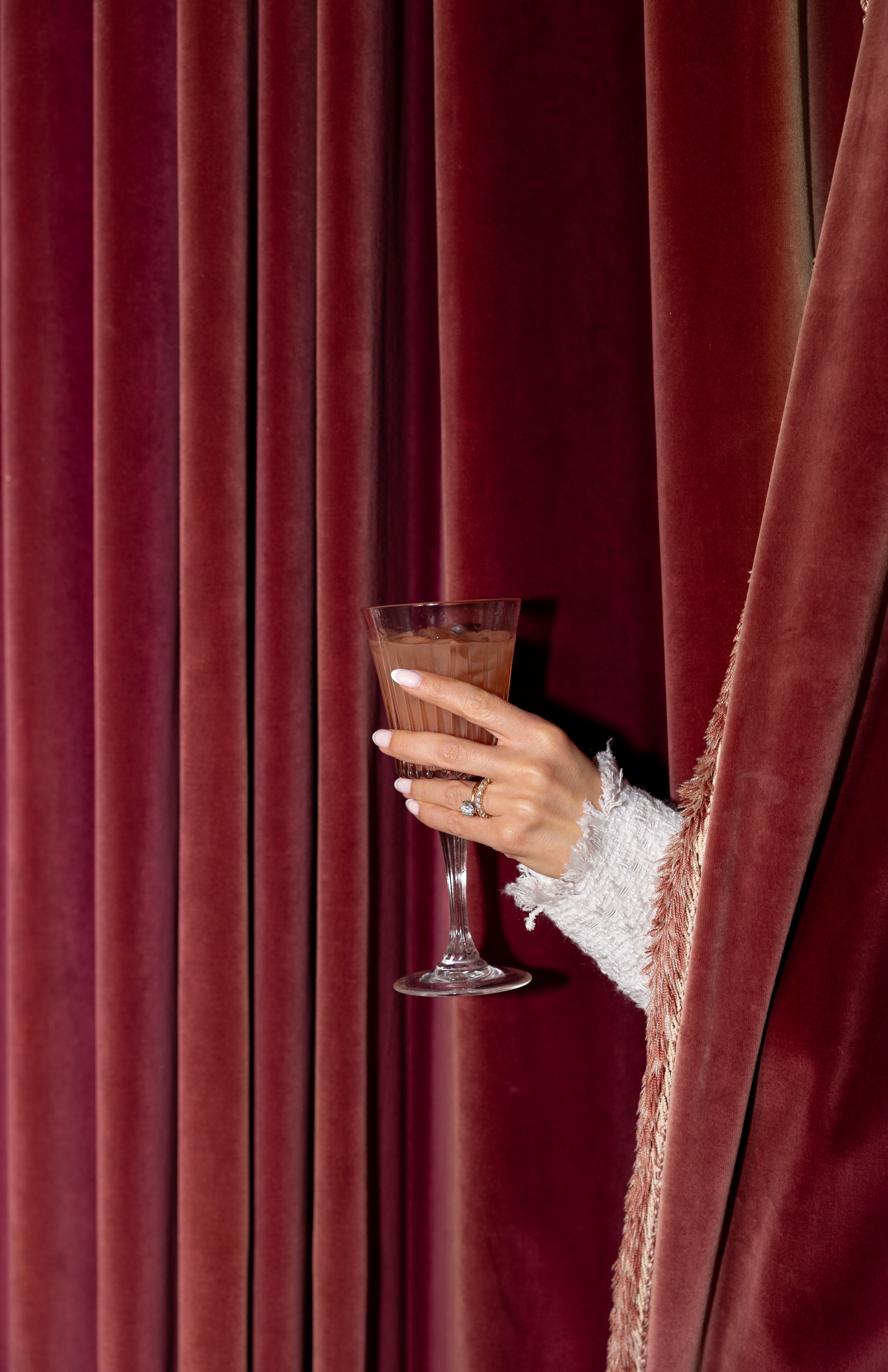 Brides hand wearing diamond ring emerges from rich burgundy velvet curtain holding elegant crystal champagne flute.