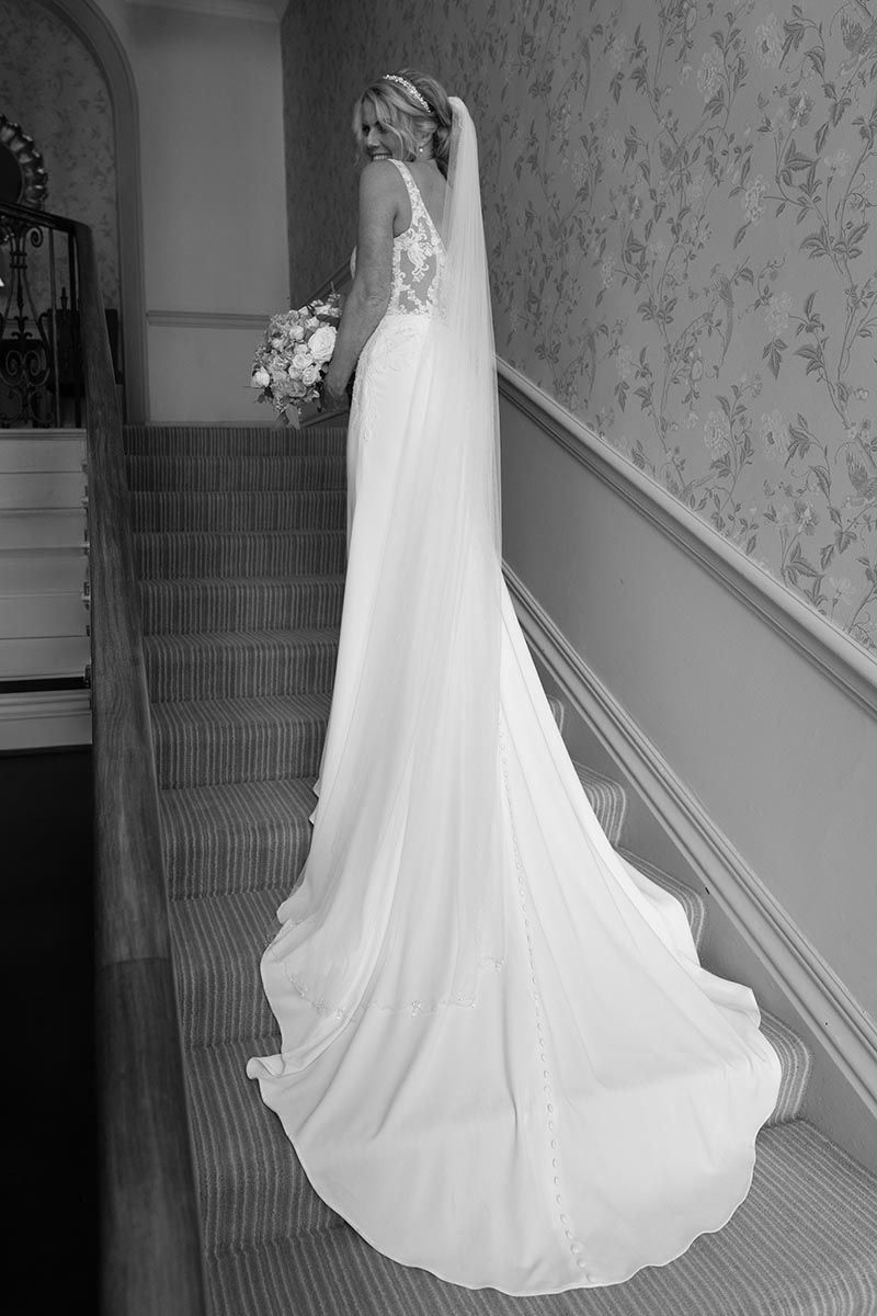 Bride in elegant white wedding gown with long train and veil stands on the grand staircase at the Belsfield Hotel in Windermere