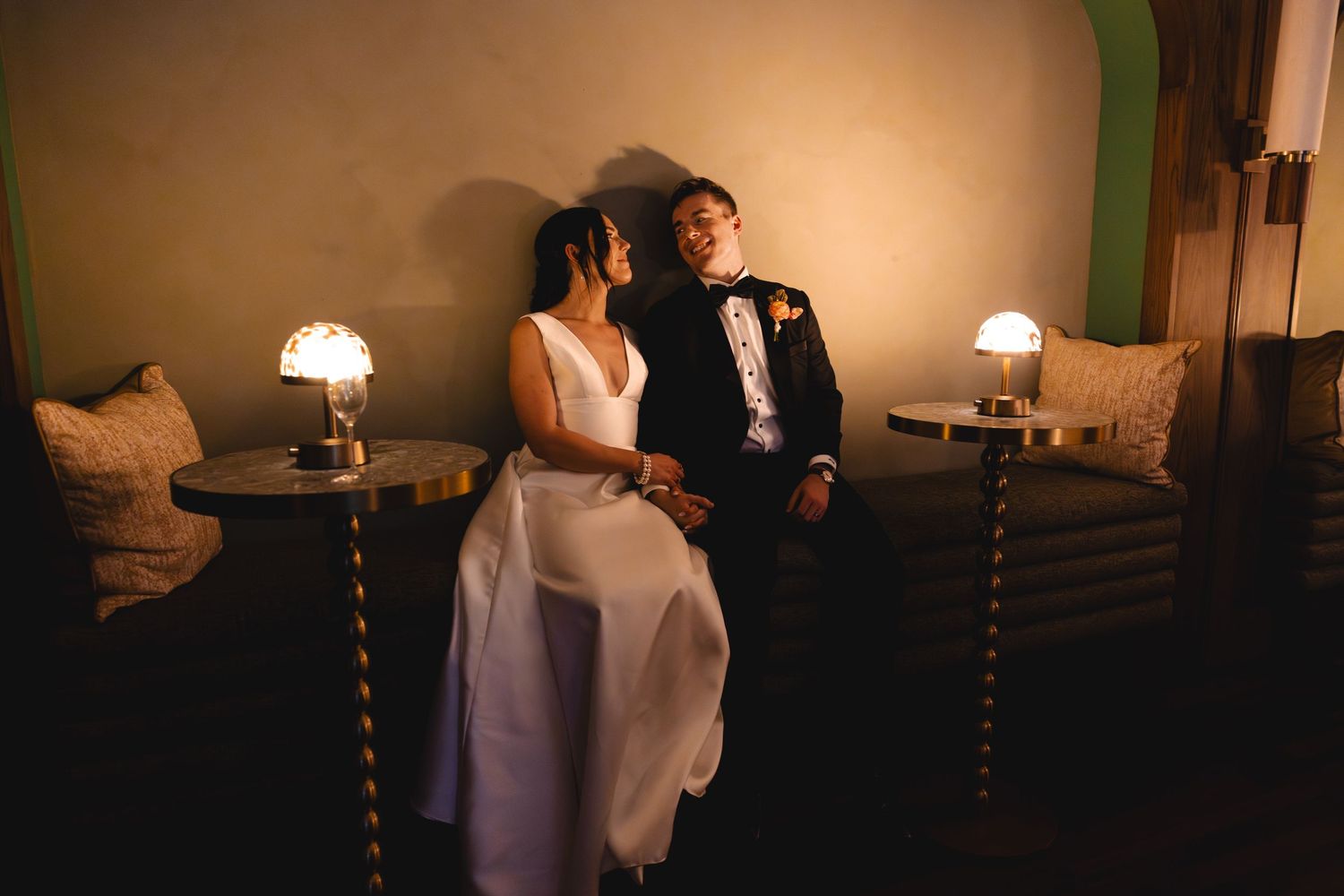 Bride in a white gown and groom in a dark suit sit close together on a cushioned bench between glowing lamps in a softly lit, elegant room.