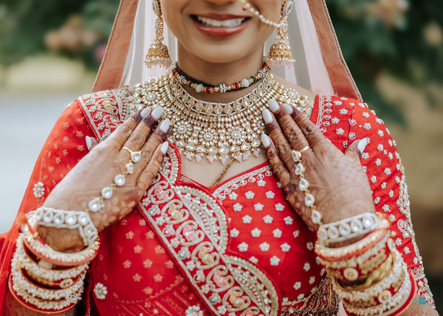 Smiling bride in a red and gold embroidered lehenga with kundan jewelry, a statement necklace, bangles, and intricate henna on her hands.