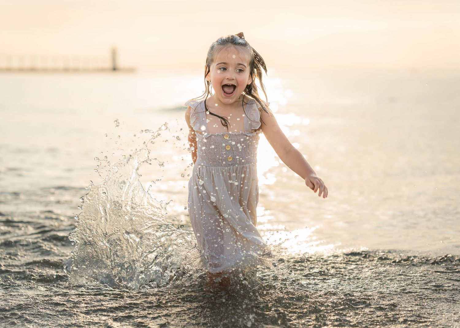 A joyful young girl in a white dress splashing through shallow ocean waves at sunset.