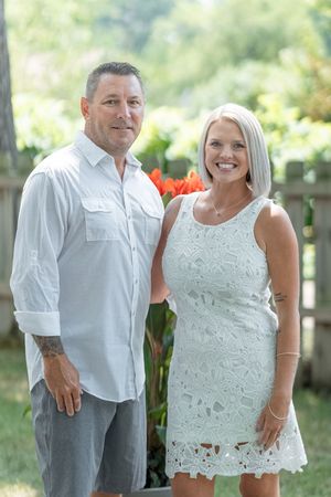 A smiling couple in white outfits poses outdoors in a lush garden setting with vibrant orange flowers in the background.