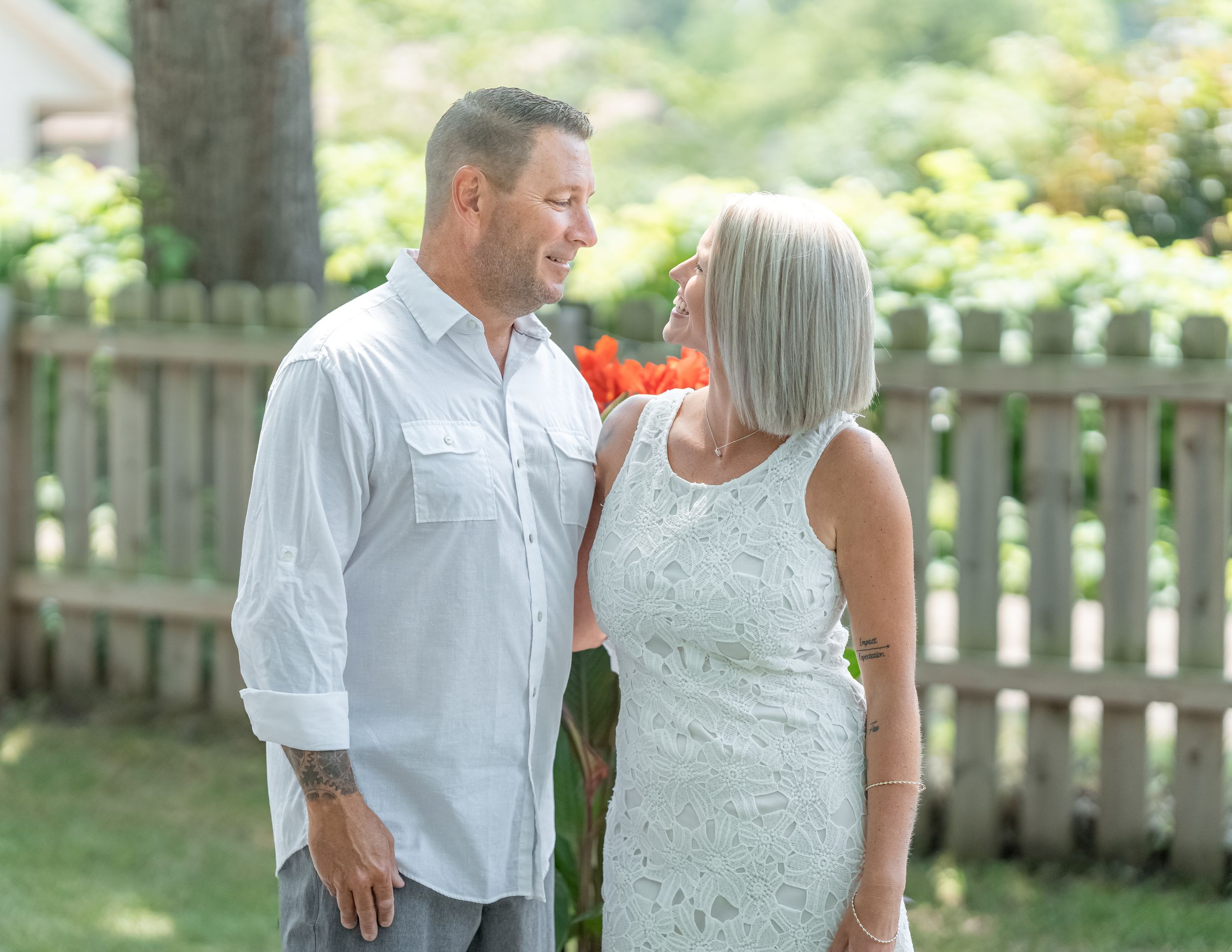 Couple smiling at each other outdoors, man in white shirt and woman in lace dress, with a wooden fence background.