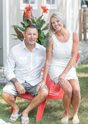 A smiling couple in white outfits sitting on a red Adirondack chair outdoors on a sunny day.