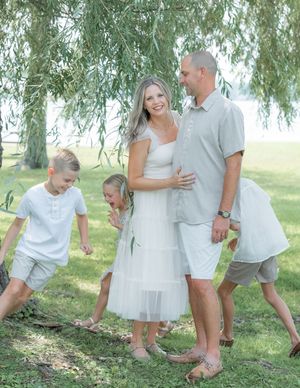 Happy family of five posing outdoors under willow tree, dressed in white and grey coordinated outfits.