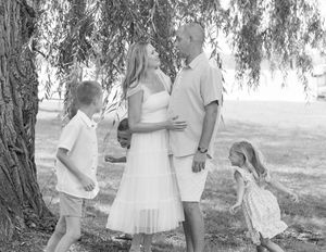 Black and white family photo under willow tree with parents and two young children in white outfits outdoors.