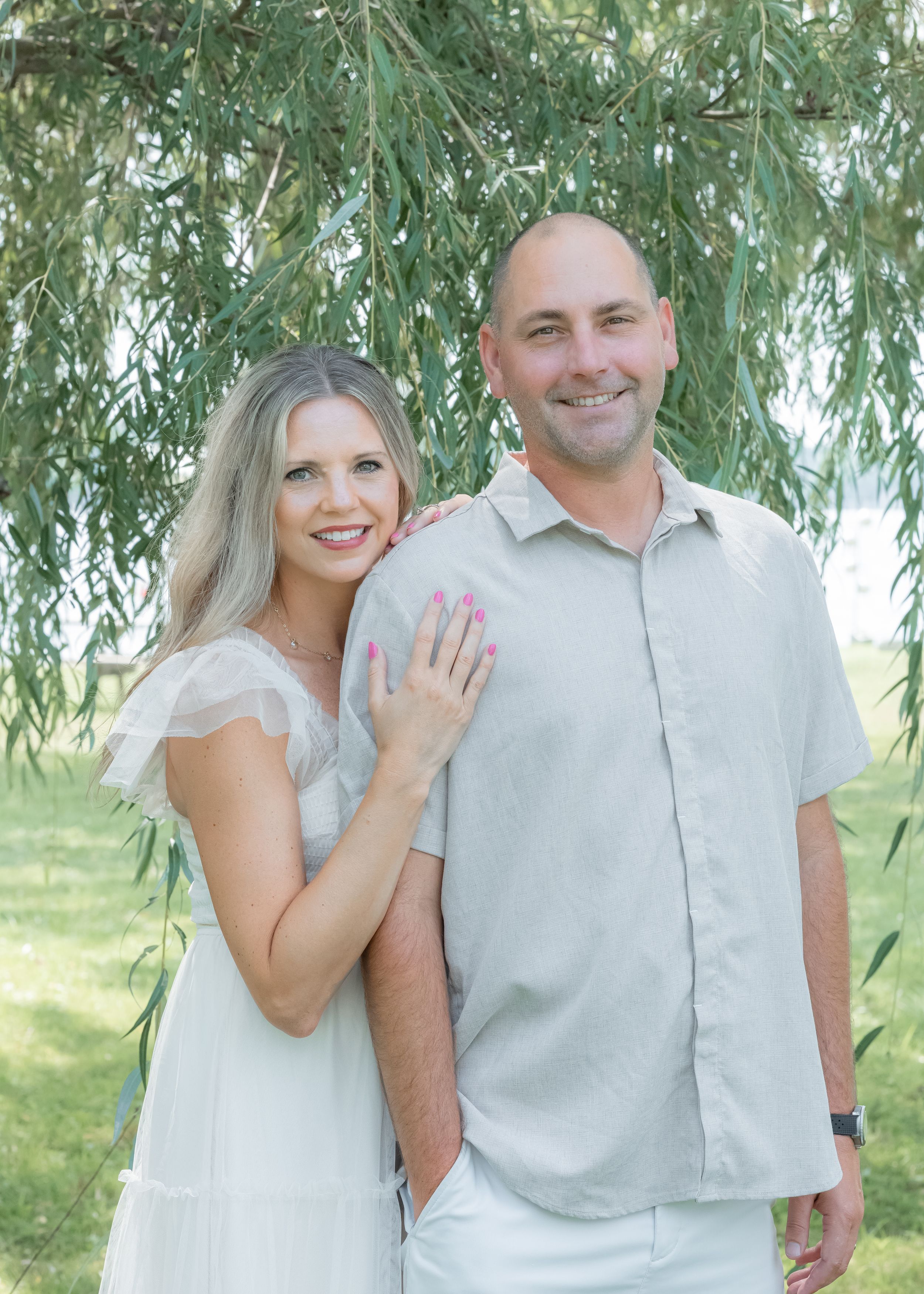 Smiling couple posing outdoors under a weeping willow tree, woman in white dress showing engagement ring.